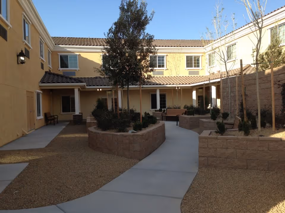 Outdoor courtyard area of a senior living facility with a paved walkway, raised garden beds, small trees, benches, and a two-story yellow building surrounding the courtyard under a clear blue sky.