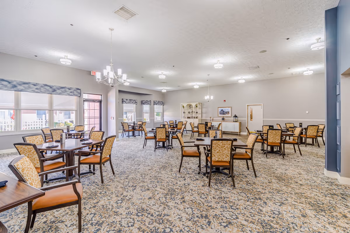 A spacious dining room with multiple square tables and cushioned chairs arranged neatly. The room has large windows with patterned valances allowing natural light to enter. The walls are painted light gray, and the carpet has a floral pattern. There are chandeliers and ceiling lights providing illumination, and a white cabinet with decorative items is visible against the far wall.