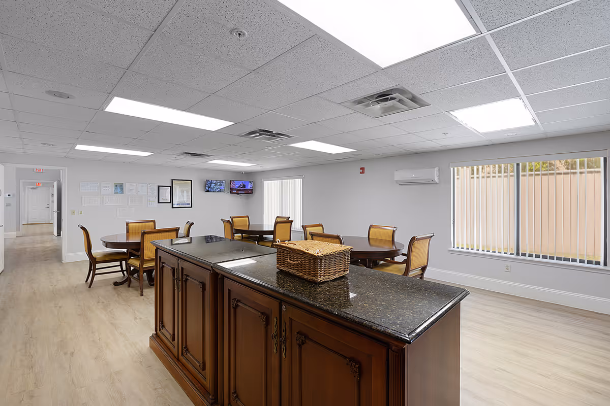 A bright and spacious dining area in a senior living facility with wooden tables and chairs arranged neatly. The room features a large wooden island with a granite countertop and a wicker basket on top. There are two wall-mounted televisions and framed documents on the wall. Large windows with vertical blinds allow natural light to enter the room.