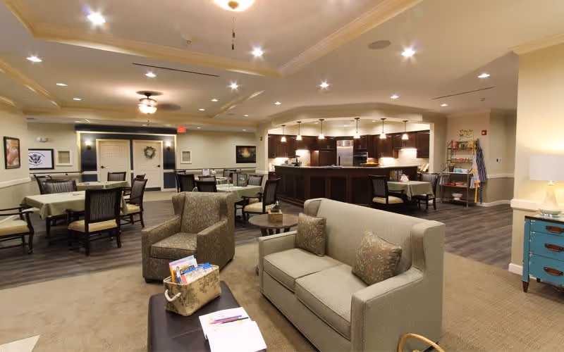 Spacious senior living common area with sofas, armchairs, dining tables, and a serving bar in the background.