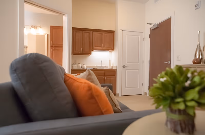 Interior view of a senior living facility room showing a gray couch with orange and beige pillows in the foreground, a small round table with a green plant, a kitchenette with wooden cabinets and a sink, a closed white door, and a brown entrance door.