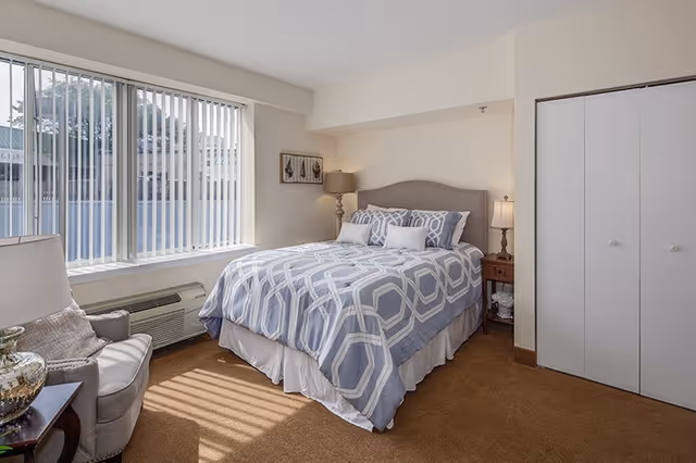 A bedroom with a large window with vertical blinds letting in natural light. The room features a bed with a gray and white patterned comforter and multiple pillows. There is a nightstand with a lamp on each side of the bed, a gray armchair with a pillow, and a closet with white double doors. The walls are painted light beige and the carpet is brown.