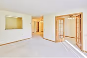 Bright and empty living room area with beige carpet and white walls. There is a small pass-through window to the kitchen on the left wall, and double glass-paneled French doors on the right leading to a dining room with a chandelier. Sunlight streams in through the glass doors, casting shadows on the floor.