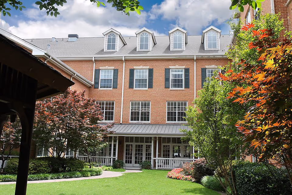 A red brick multi-story building with white-framed windows and dormer windows on the roof, surrounded by a well-maintained garden with green grass, colorful trees, and shrubs under a partly cloudy sky.