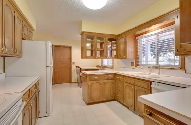 A spacious kitchen with wooden cabinets, white countertops, a white refrigerator, a white stove, and a dishwasher. There is a window with blinds above the sink, and a small dining area with chairs is visible in the background near a wooden door.