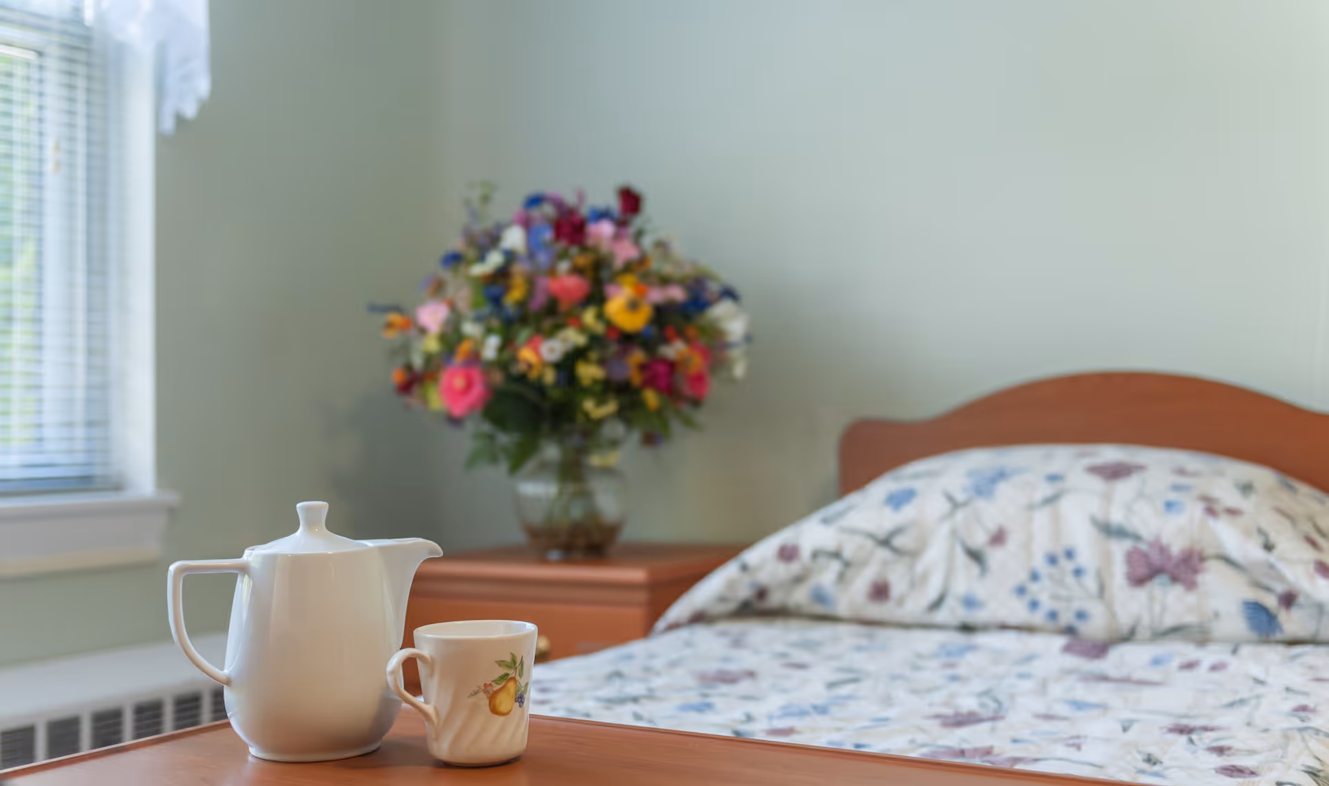 A cozy bedroom with a wooden bed featuring floral patterned bedding. On a wooden table in the foreground, there is a white teapot and a matching cup with a fruit design. In the background, a vase filled with colorful flowers sits on a nightstand next to the bed. A window with blinds allows natural light into the room.