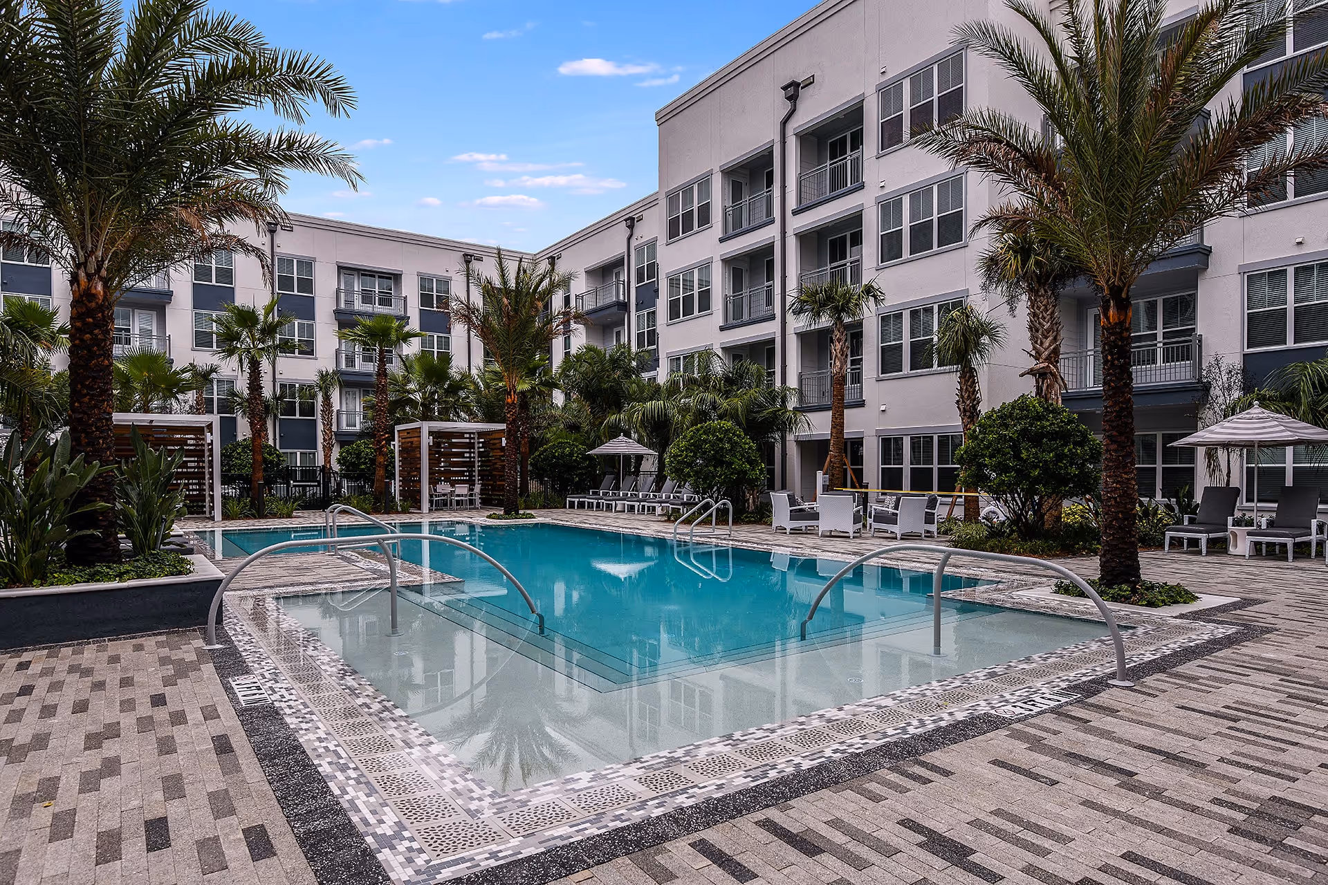 Outdoor swimming pool area surrounded by palm trees and lounge chairs with umbrellas. The pool is located in the courtyard of a modern multi-story residential building with balconies and large windows.