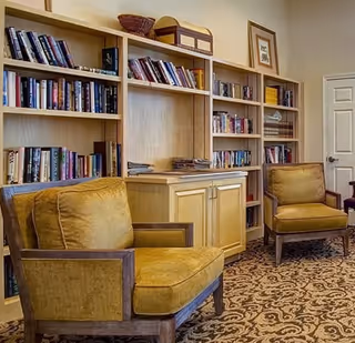A cozy reading area with two mustard yellow cushioned armchairs in front of built-in wooden bookshelves filled with books and decorative items. The room has a patterned carpet and a closed white door in the background.
