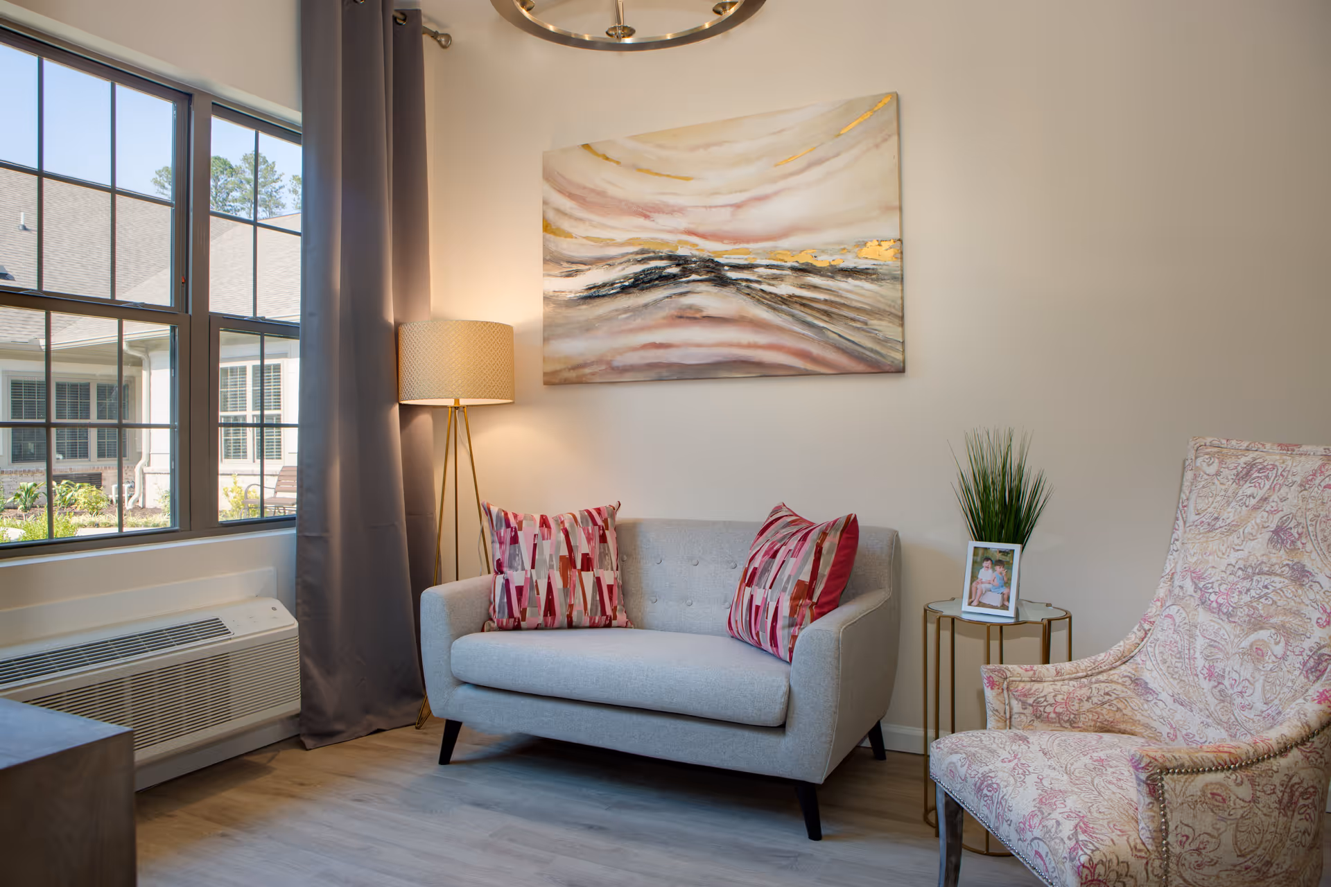 A cozy living room corner with a small gray loveseat, patterned pillows, a floor lamp, an upholstered armchair, a side table with decor, and an abstract painting above.