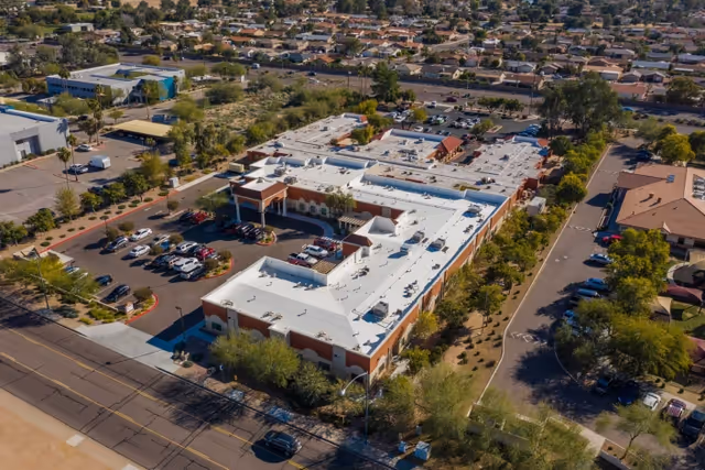 Aerial view of a large single-story building with a white roof surrounded by parking lots, trees, and roads. The building is located in a suburban area with houses and other buildings in the background.