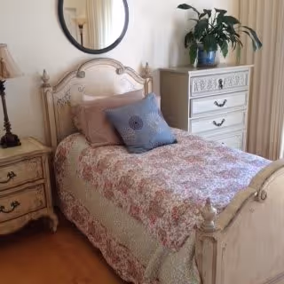 A cozy bedroom with a single bed featuring a floral quilt and two pillows, one pink and one blue. The bed has a vintage-style wooden headboard and footboard. Next to the bed is a wooden nightstand with a lamp. On the right side, there is a white dresser with decorative carvings and a large green plant on top. A round mirror hangs on the wall above the headboard.