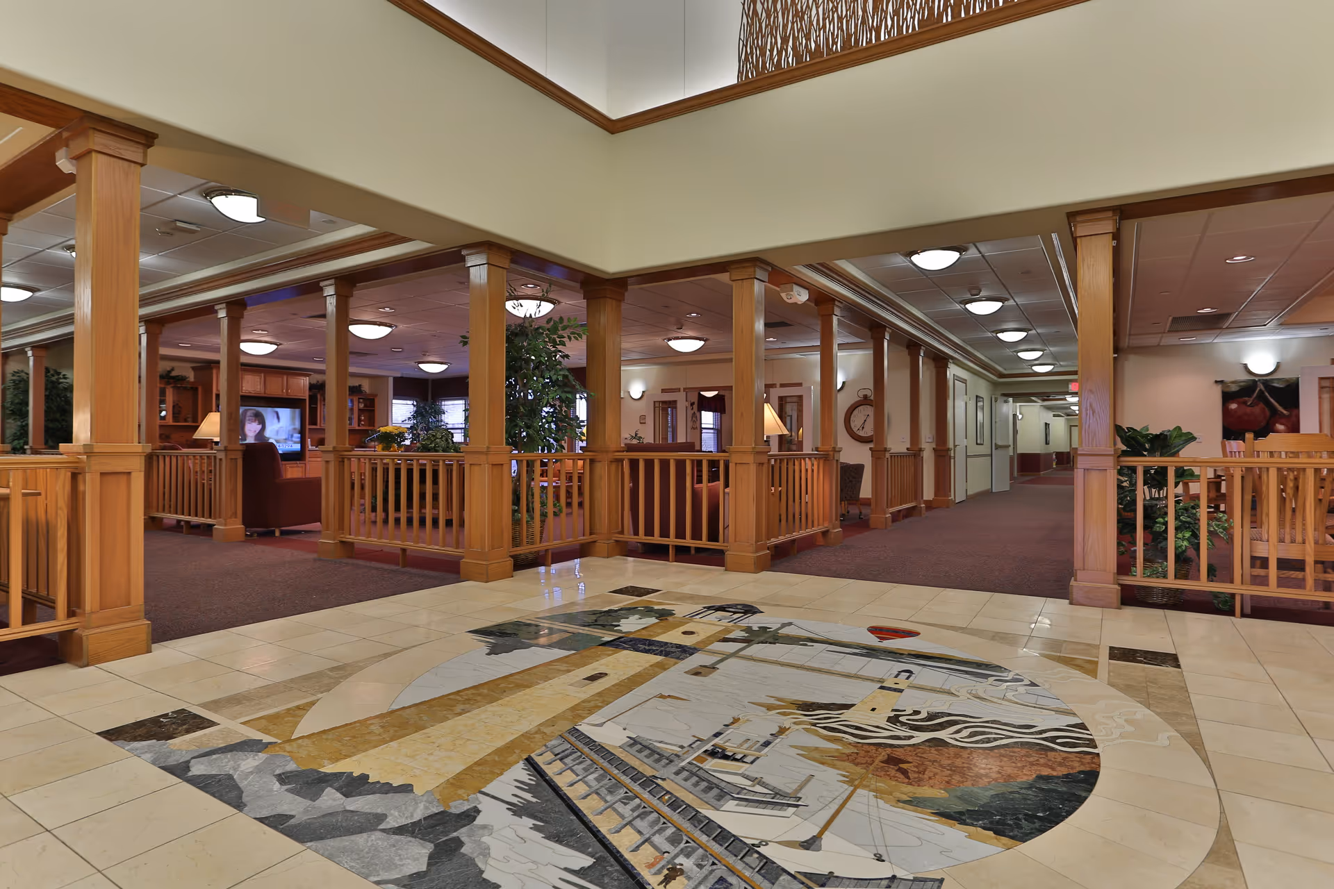 Interior view of a senior living facility lobby area with wooden columns and railings surrounding a seating area. The floor features a large decorative tile mosaic of a lighthouse and coastal scene. The space is well-lit with ceiling lights and has carpeted and tiled flooring sections.
