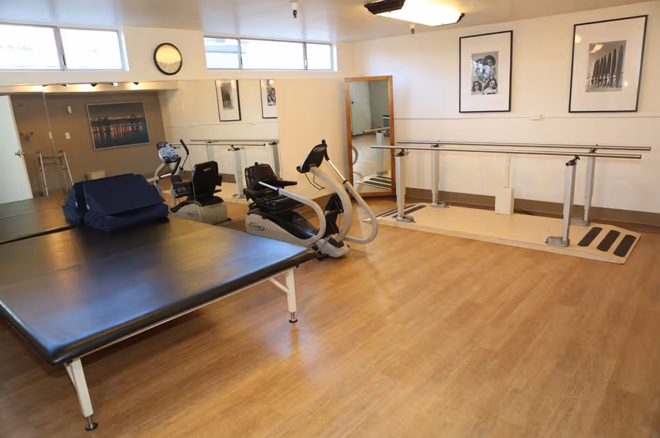 A bright therapy/fitness room with exercise equipment including recumbent bikes, parallel bars, and a large padded treatment table.