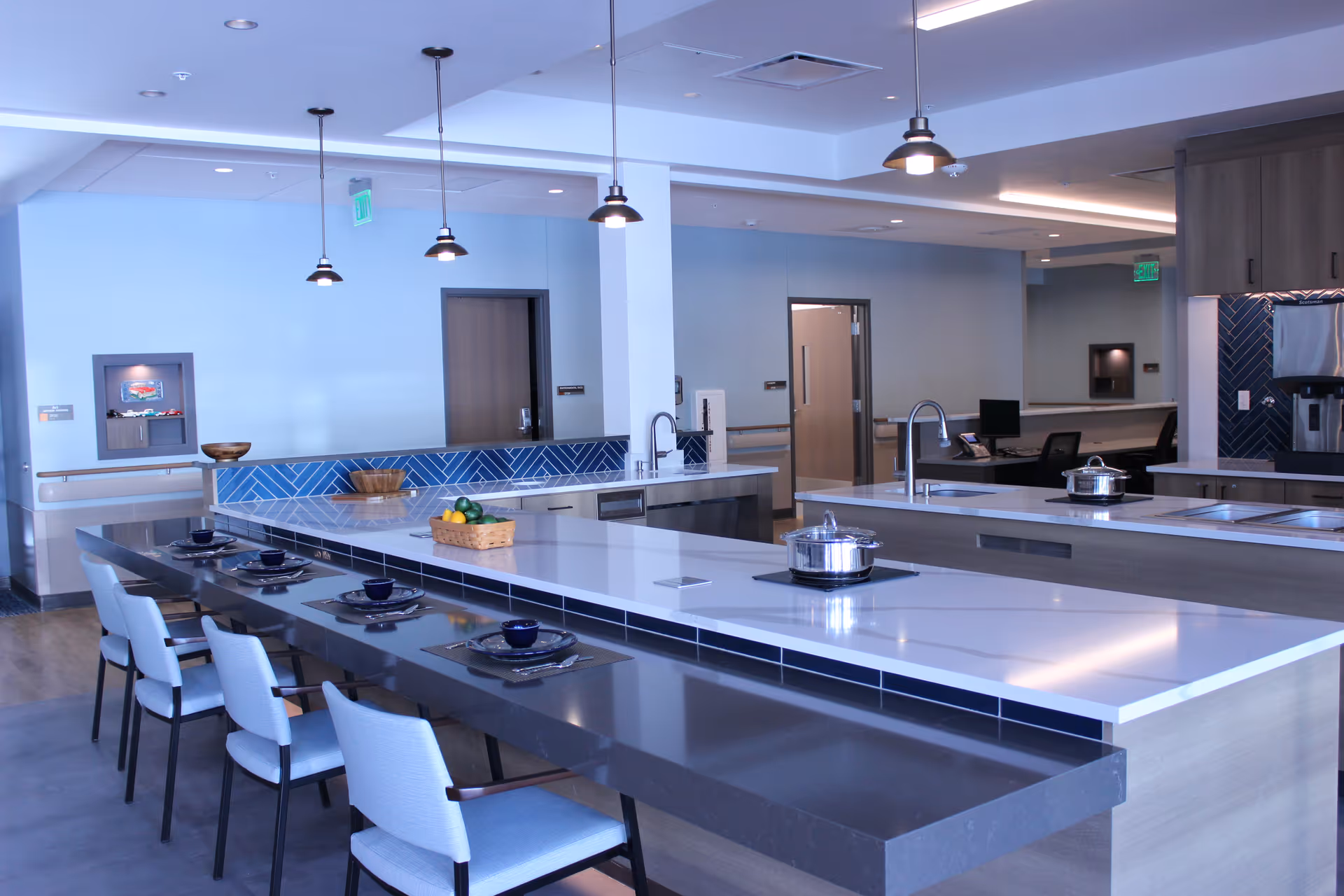 Modern kitchen area with a long island countertop featuring a white marble surface and a dark extension with six chairs set with plates and bowls. The kitchen has multiple sinks, stainless steel appliances, blue tiled backsplash, and pendant lights hanging from the ceiling. The room has light blue walls and wooden doors in the background.