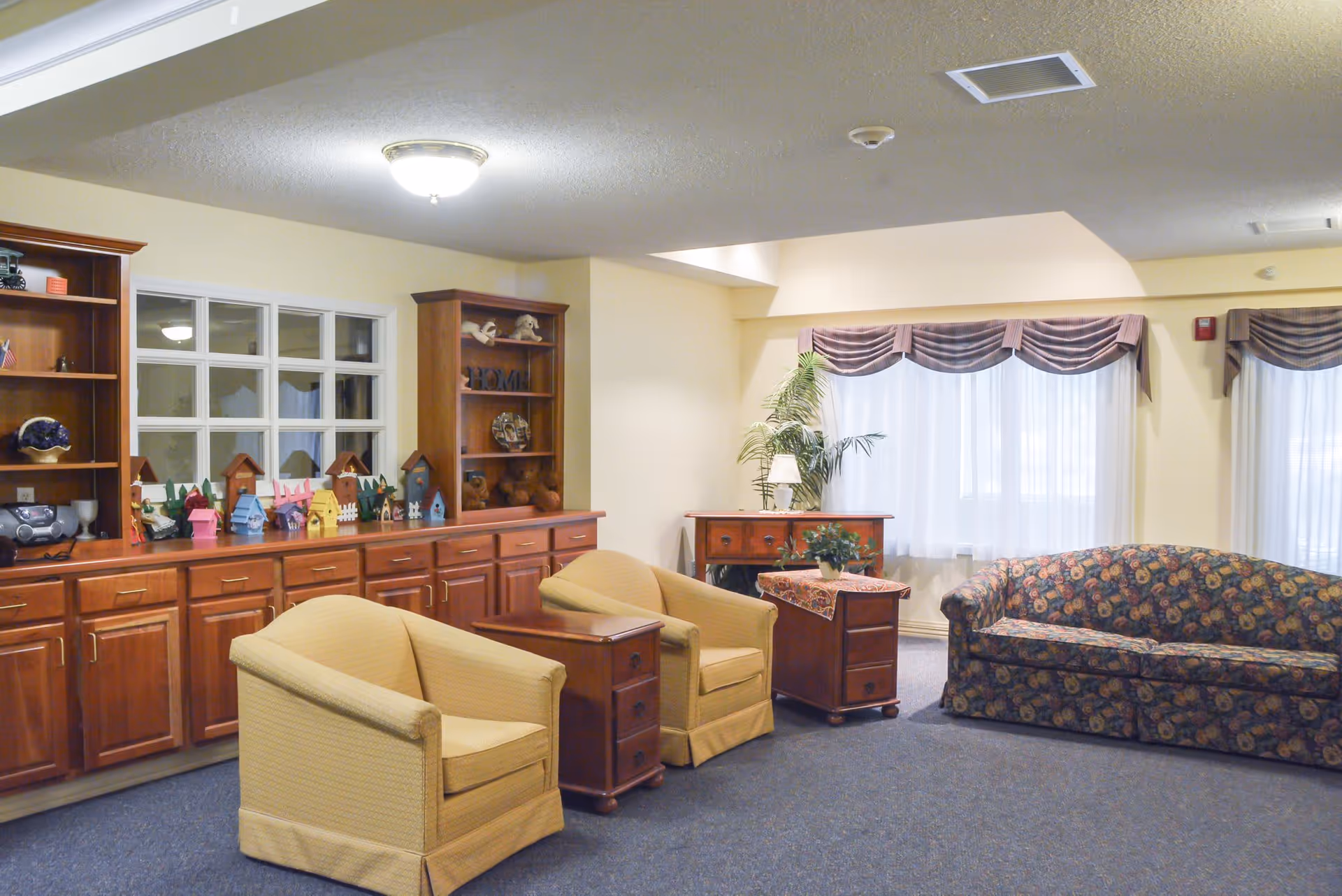 A cozy common room with upholstered armchairs, a patterned sofa, wooden cabinets and side tables under soft ceiling lighting.