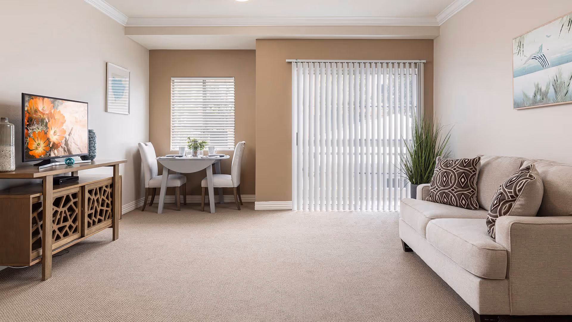 A bright living room with beige carpet and walls, featuring a beige sofa with patterned cushions on the right, a wooden TV stand with a flat-screen TV displaying orange flowers on the left, a small round dining table with two chairs near a window with blinds, and vertical blinds covering a sliding glass door in the background.