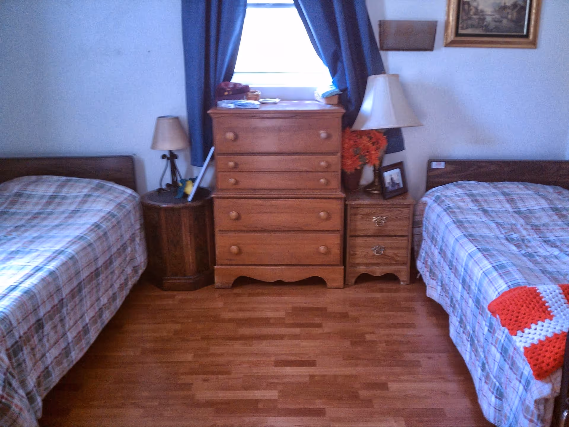 A bedroom with two single beds on either side of a wooden dresser. Each bed has a plaid bedspread, and there are two bedside tables with lamps. A window with blue curtains is above the dresser, and a framed picture and a vase with flowers are on the right bedside table.