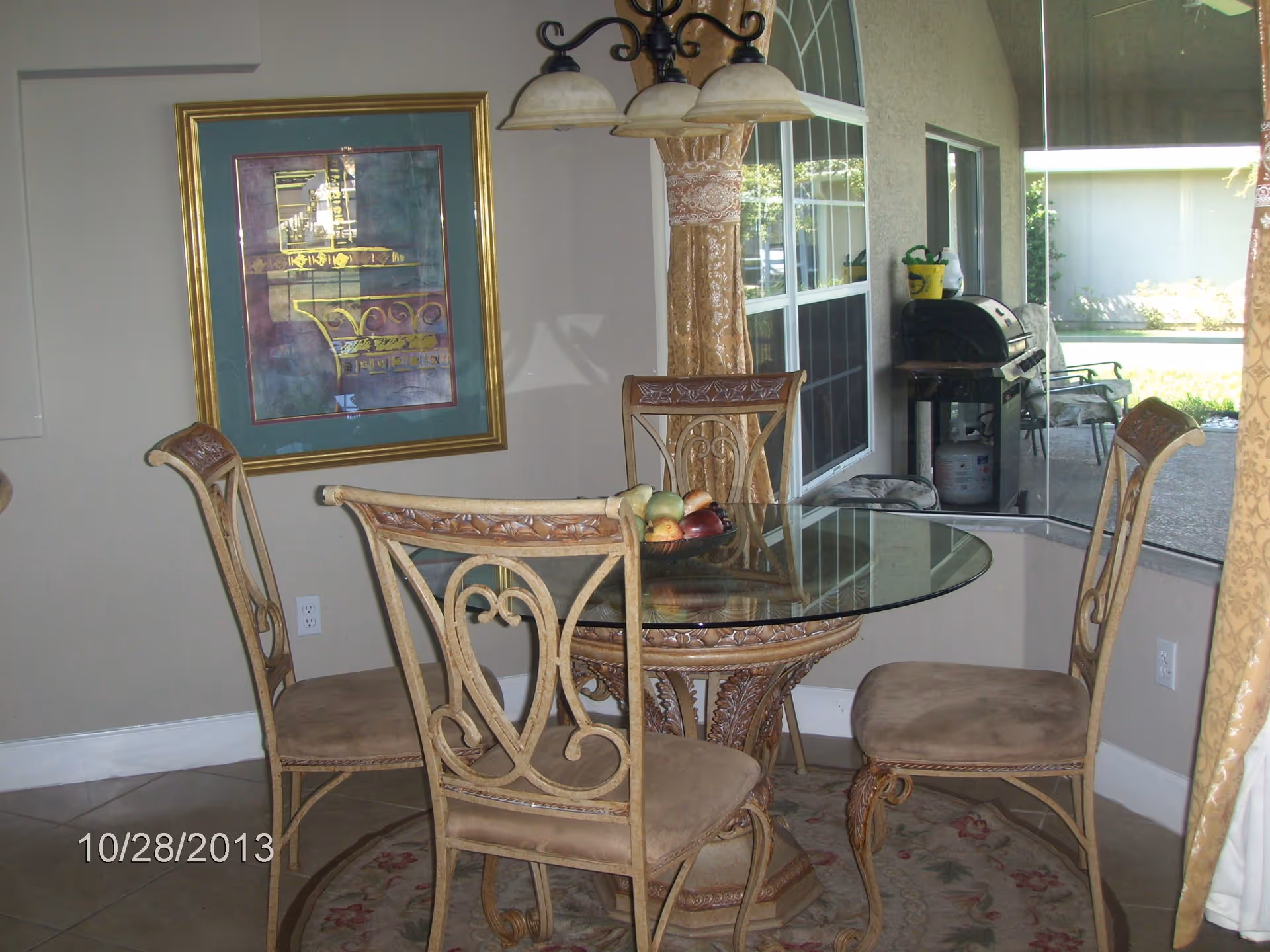 A dining area with a round glass-top table and four ornate chairs with cushioned seats. A bowl of fruit is placed on the table. Behind the table is a large window showing an outdoor patio with a grill and chairs. There is a framed artwork on the wall and a hanging light fixture above the table.
