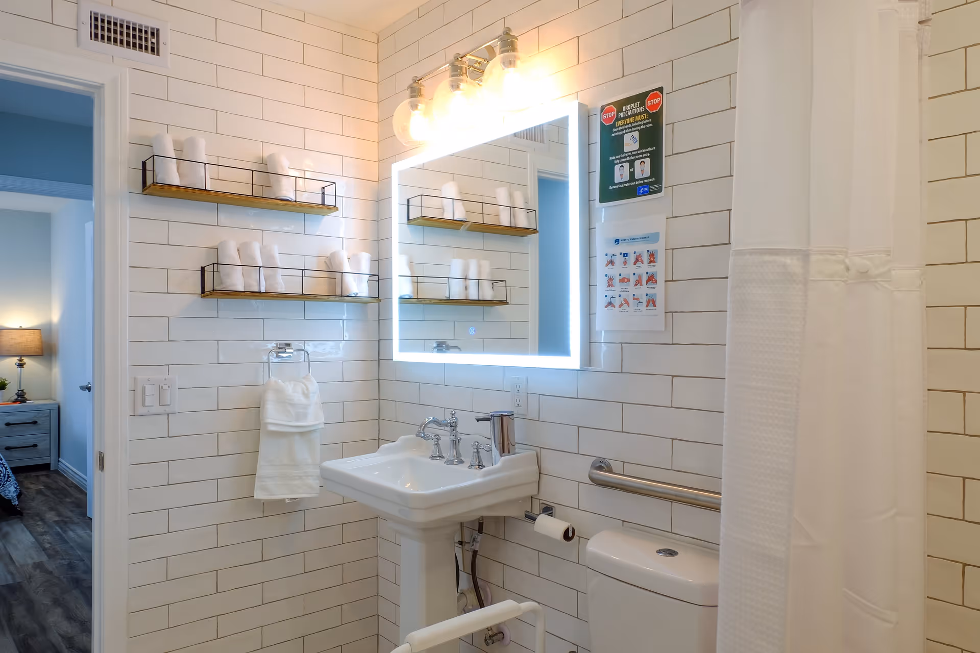 A clean, modern bathroom with white subway tile walls, a pedestal sink, a backlit mirror, and a toilet with a grab bar. Two wooden shelves hold neatly rolled white towels. A white shower curtain is partially visible on the right. A doorway leads to a bedroom with a nightstand and lamp.