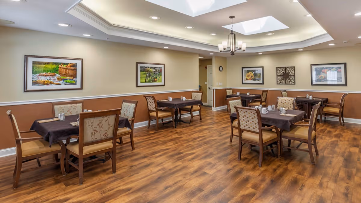 A dining room with several wooden tables covered with dark tablecloths and surrounded by cushioned chairs. The room has wood flooring, beige walls with a brown wainscot, and framed pictures hanging on the walls. The ceiling features recessed lighting and two skylights, along with a decorative chandelier.