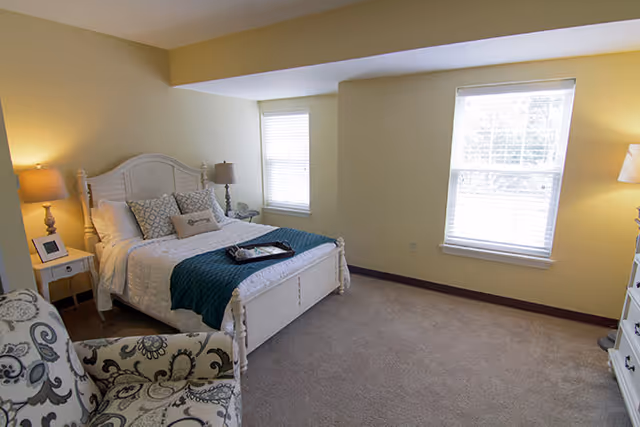 Bright bedroom with a white bed, bedside tables and lamps, a patterned armchair, and two windows.