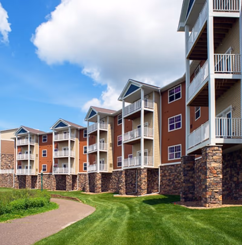 Exterior view of a multi-story senior living facility building with balconies, stone pillars, and a well-maintained lawn and walking path under a partly cloudy blue sky.