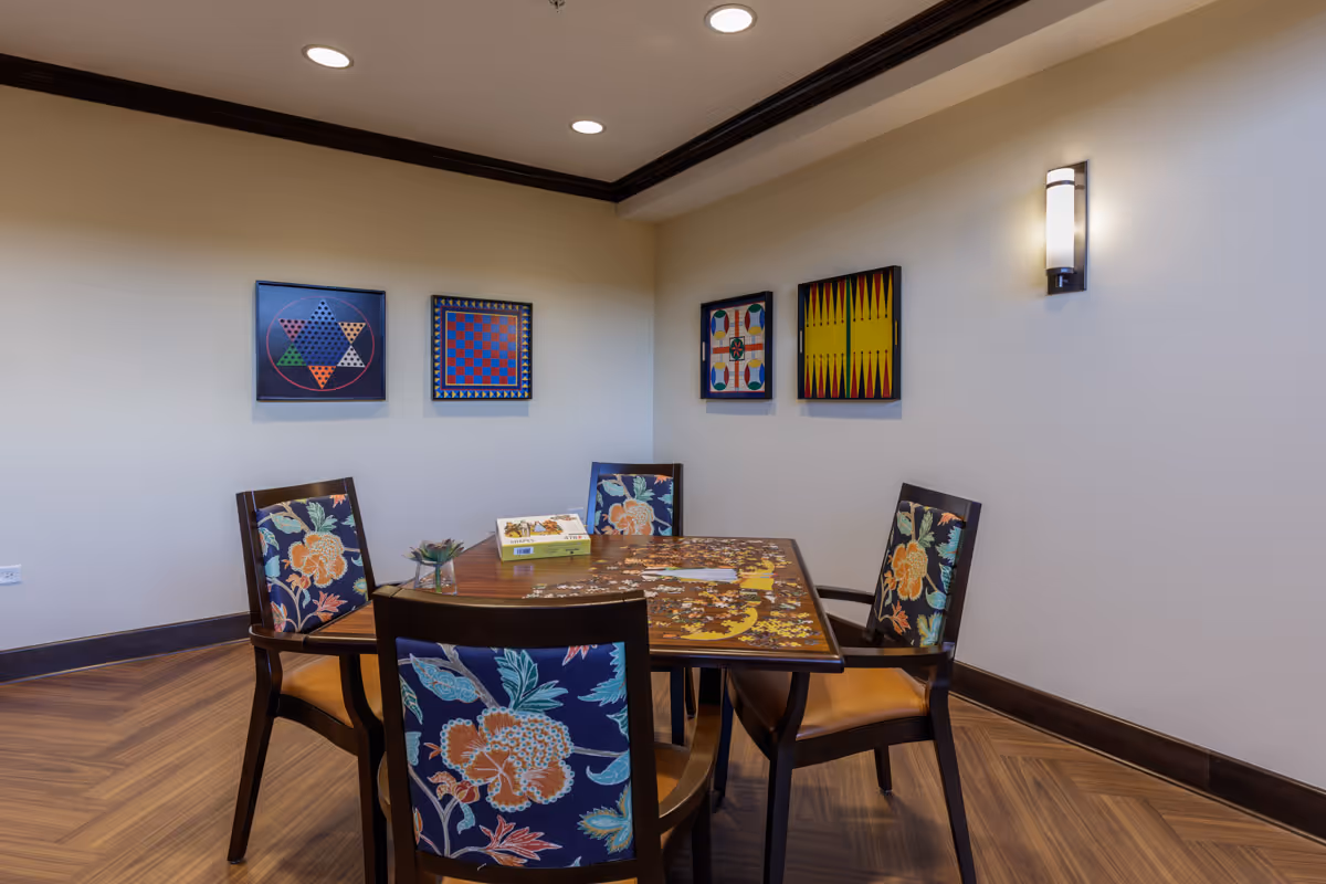 A small game room with a square wooden table and four chairs with floral upholstery. The table has a partially completed jigsaw puzzle and a box on it. The walls are decorated with four framed game boards including checkers and backgammon. The room has wooden flooring, recessed ceiling lights, and a wall-mounted light fixture.