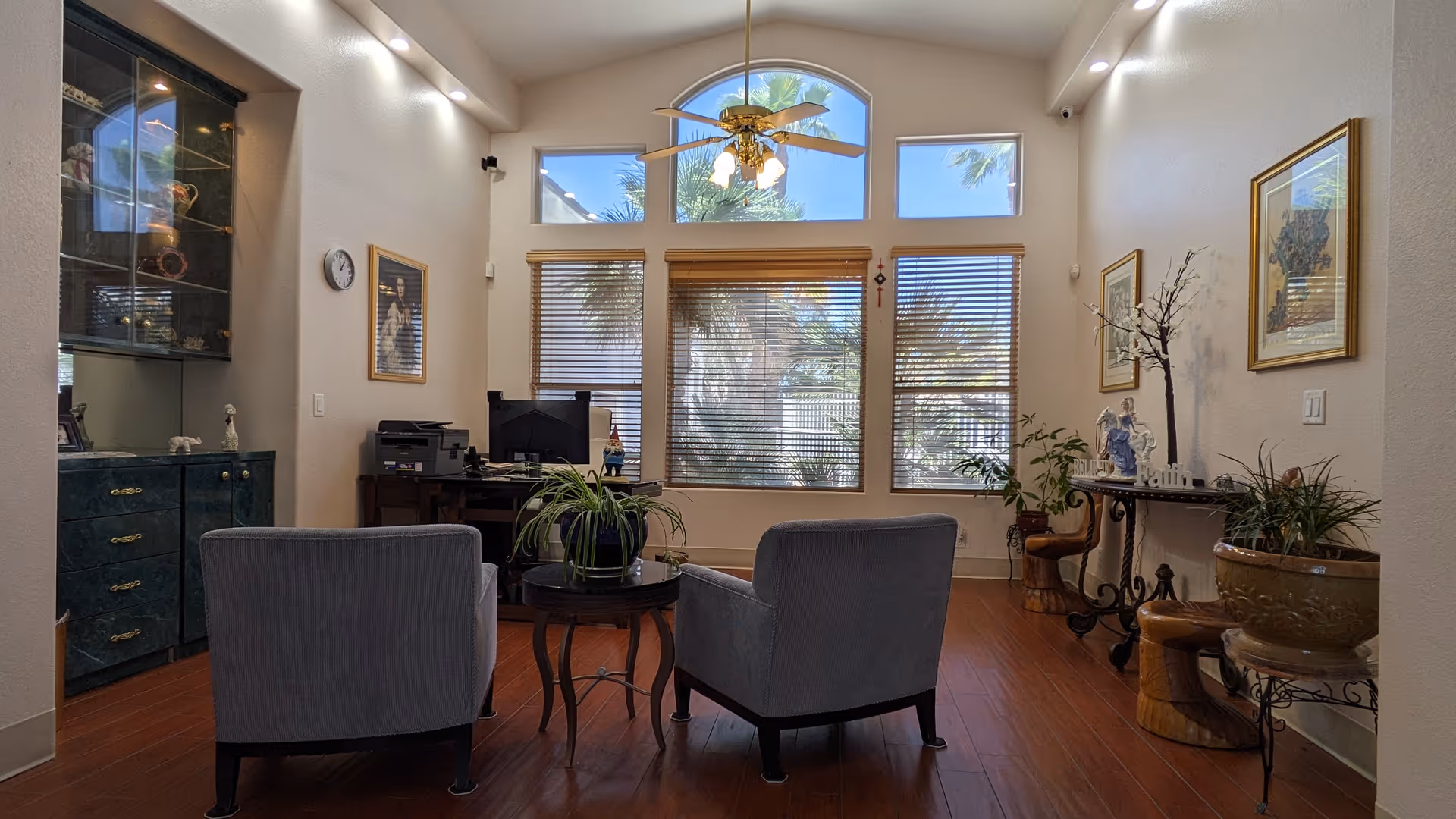 A cozy living room area with two gray armchairs facing a small round table with a potted plant. The room features large windows with wooden blinds, allowing natural light to brighten the space. There is a ceiling fan with lights, a dark green cabinet with glass doors on the left, a desk with a computer and printer, and various decorative plants and framed pictures on the walls.