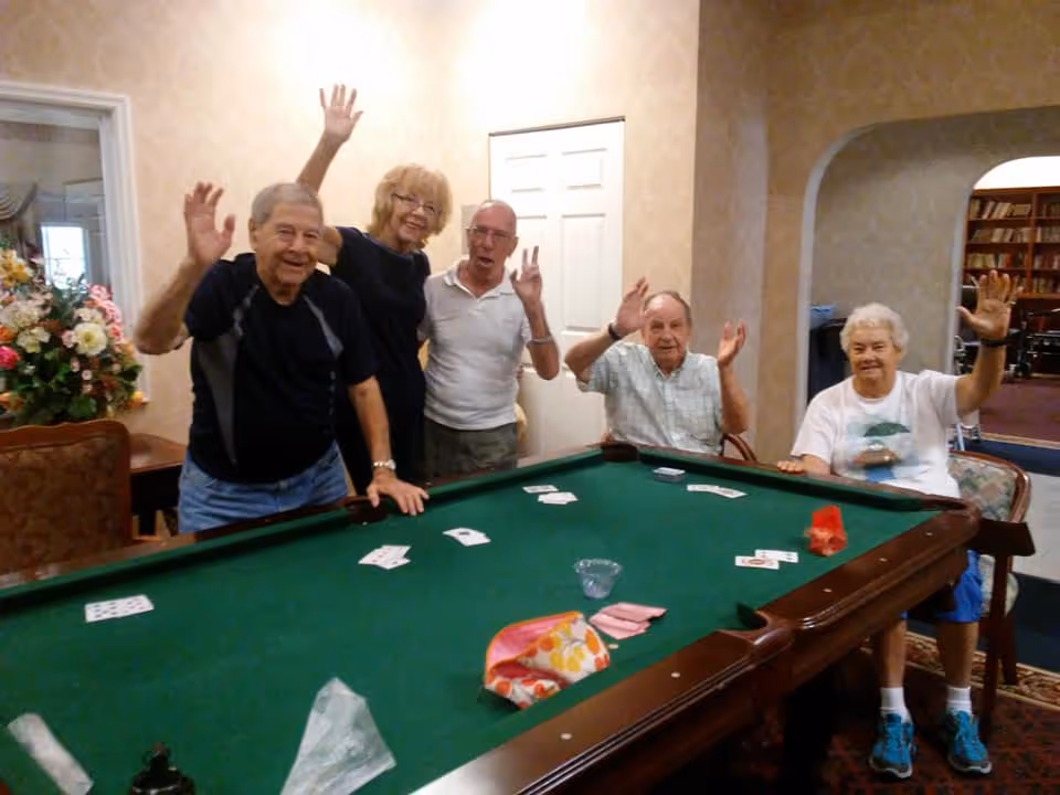 Five elderly people smiling and waving while standing and sitting around a green pool table with playing cards scattered on it in a cozy room with floral decorations and bookshelves in the background.