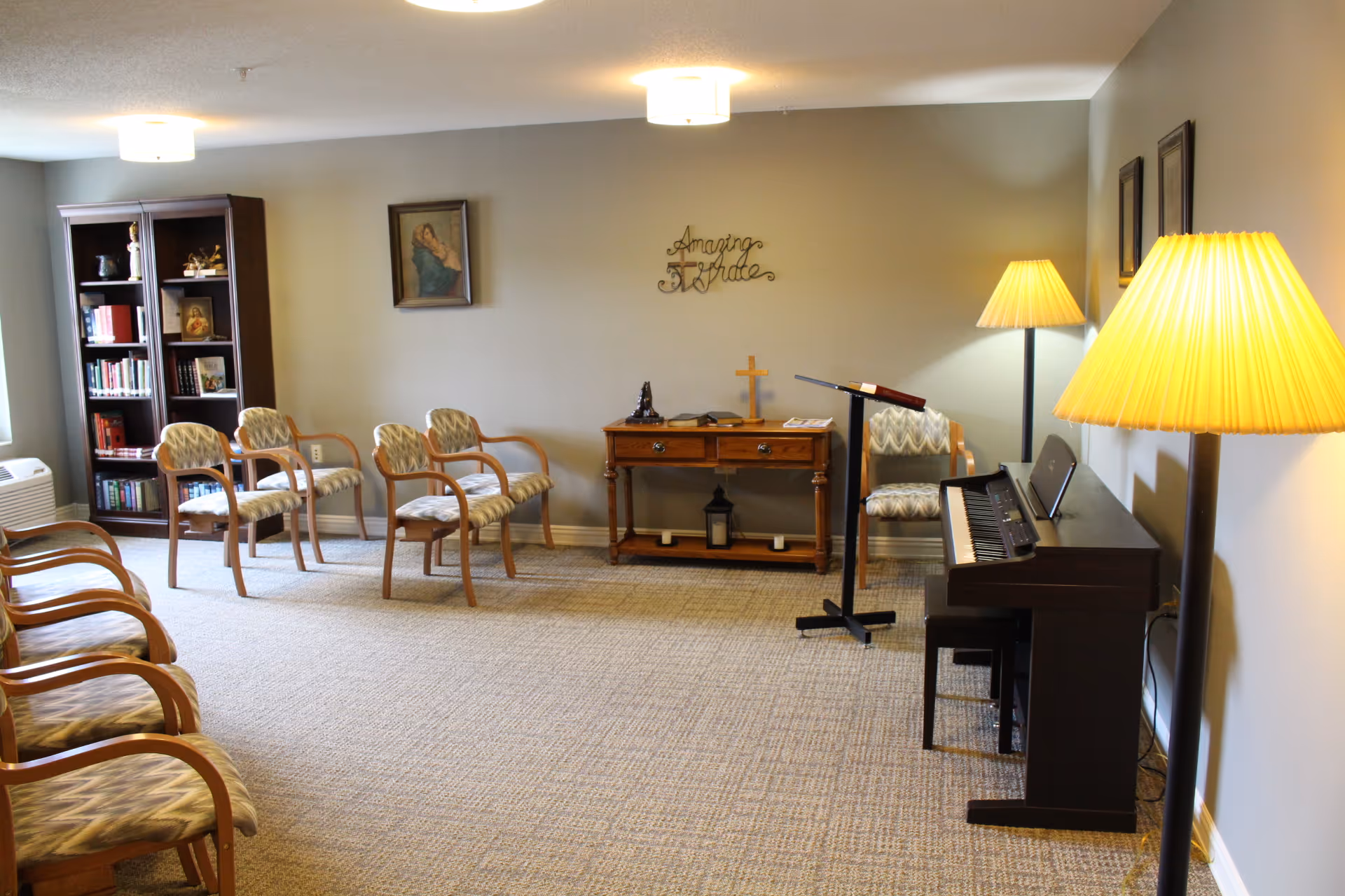 A quiet room with several wooden chairs arranged in rows facing a small wooden table with a cross and books on it. There is a piano with a music stand and a chair next to it on the right side of the room. Two floor lamps with yellow lampshades provide warm lighting. A bookshelf filled with books is against the left wall, and two framed pictures hang on the walls. The wall behind the table has a decorative sign that reads 'Amazing Grace'.