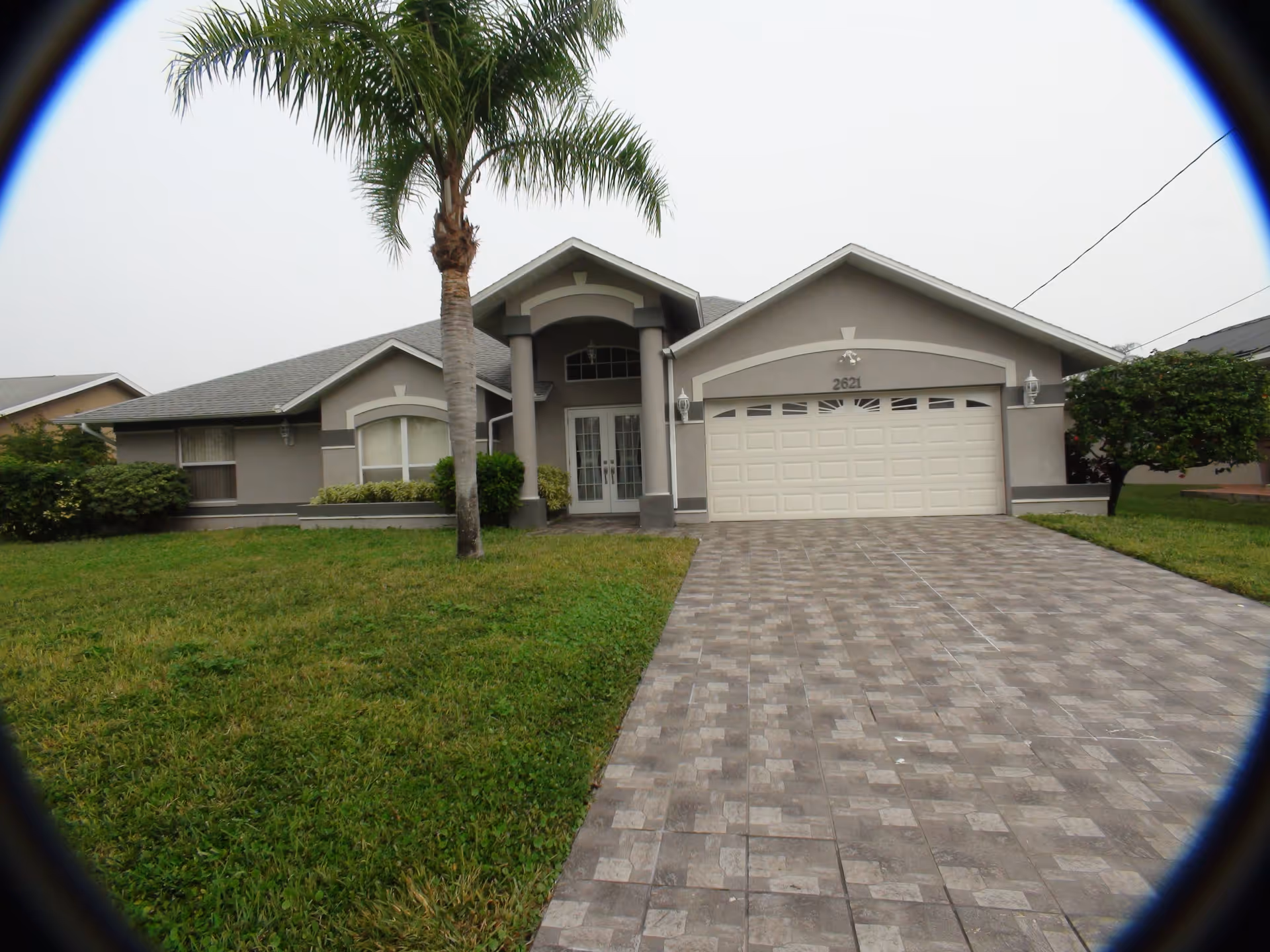 Front exterior view of a single-story residential-style building with a tiled driveway, a double garage door, a palm tree, and well-maintained grass and shrubs.