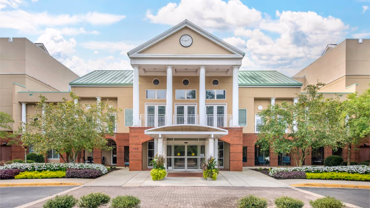Front entrance of a multi-story senior living building with tall white columns, a pediment clock, glass doors, and landscaped driveway.