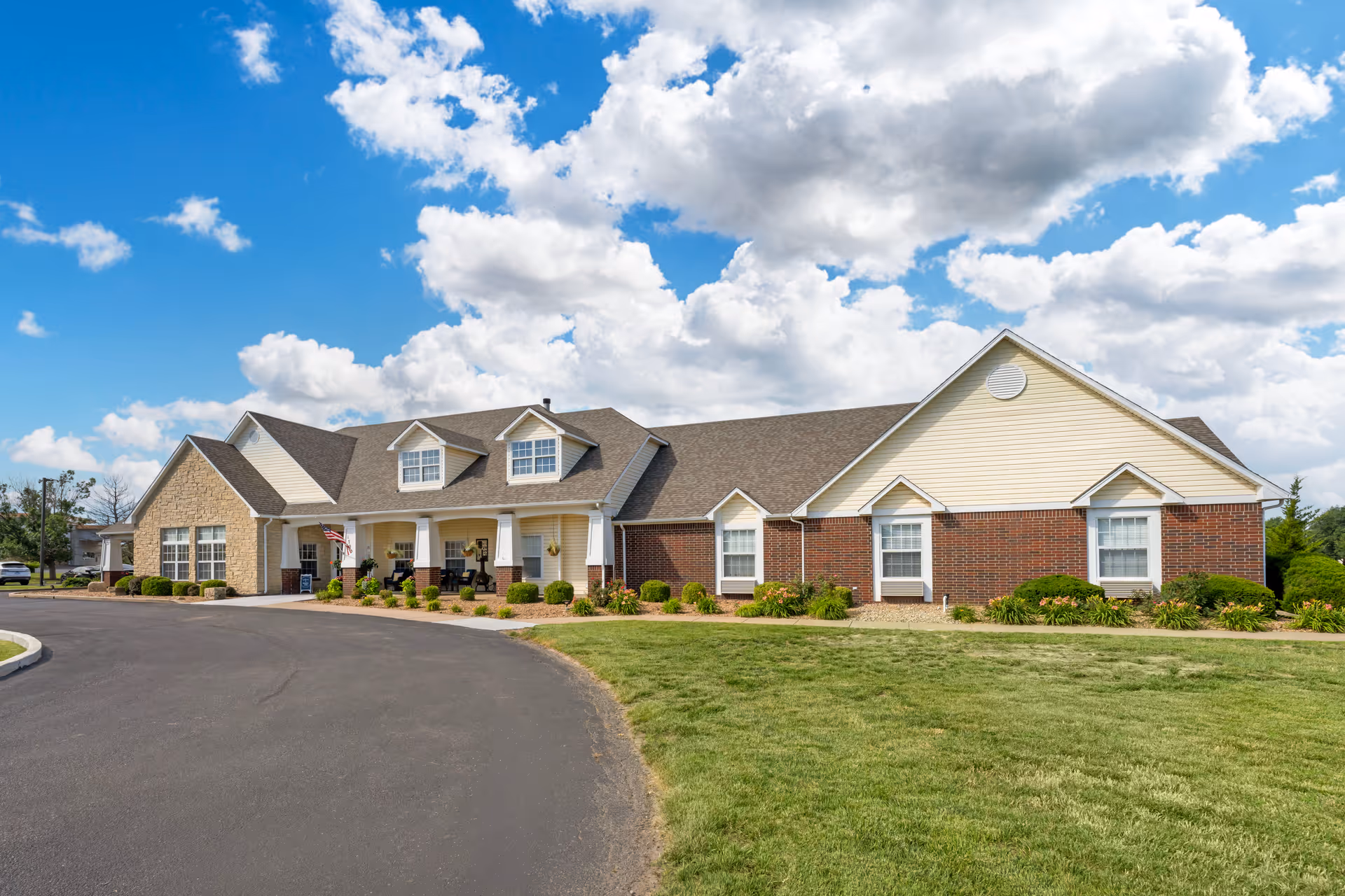 Exterior view of a single-story senior living facility building with a combination of brick and siding walls, a covered porch with seating, and a curved driveway in front. The sky is partly cloudy with bright blue patches.
