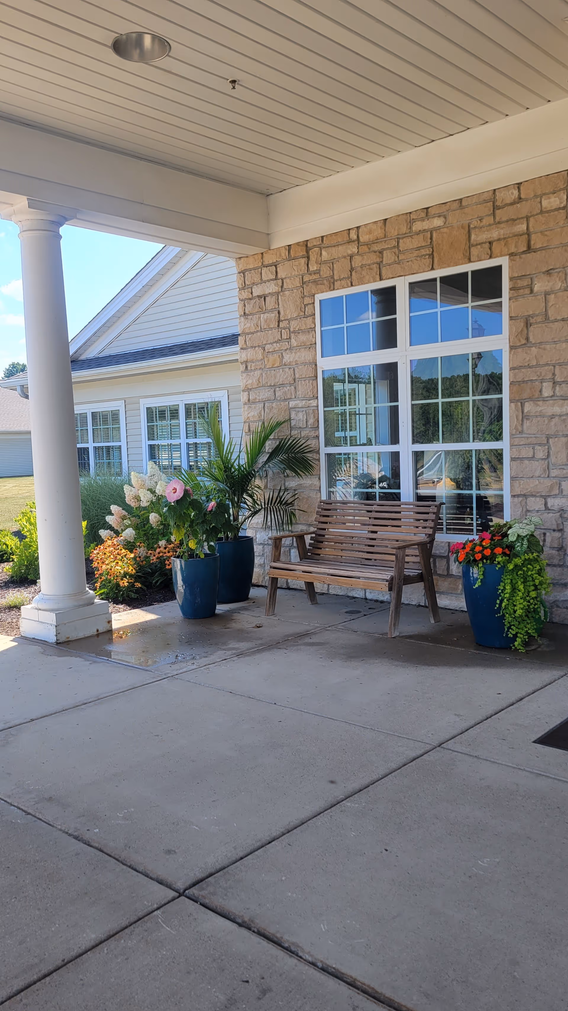 Covered outdoor seating area with a wooden bench, potted plants with flowers, and a stone wall with a large window reflecting the outdoor scenery.