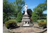 Stone statue of a robed figure on a tiered pedestal in a circular paved garden surrounded by shrubs and trees.