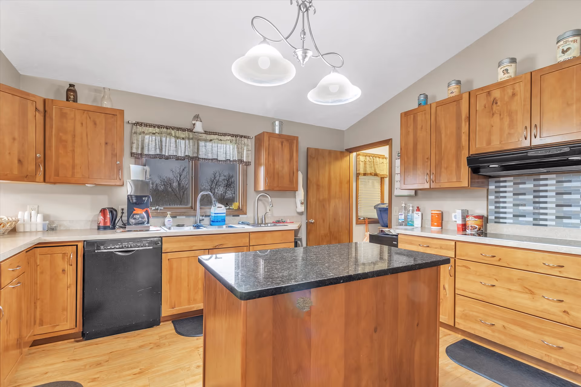 Kitchen with wooden cabinets, a central island topped with a black countertop, sink under a window, dishwasher, and overhead lighting.