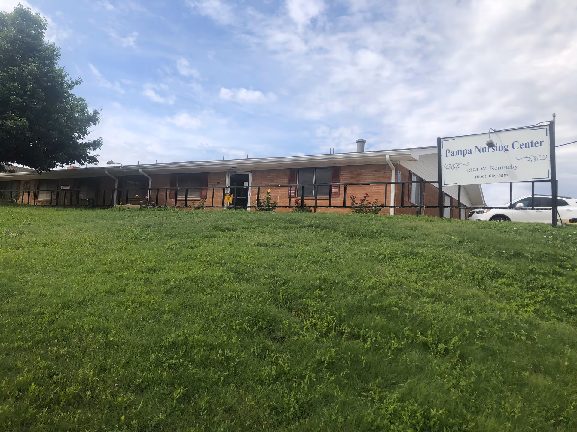 Exterior view of a single-story brick building with a sloped grassy lawn in front. A large sign near the building reads 'Pampa Nursing Center' with the address 1321 W. Kentucky and a phone number. A white vehicle is parked near the building, and a tree is visible on the left side under a partly cloudy sky.