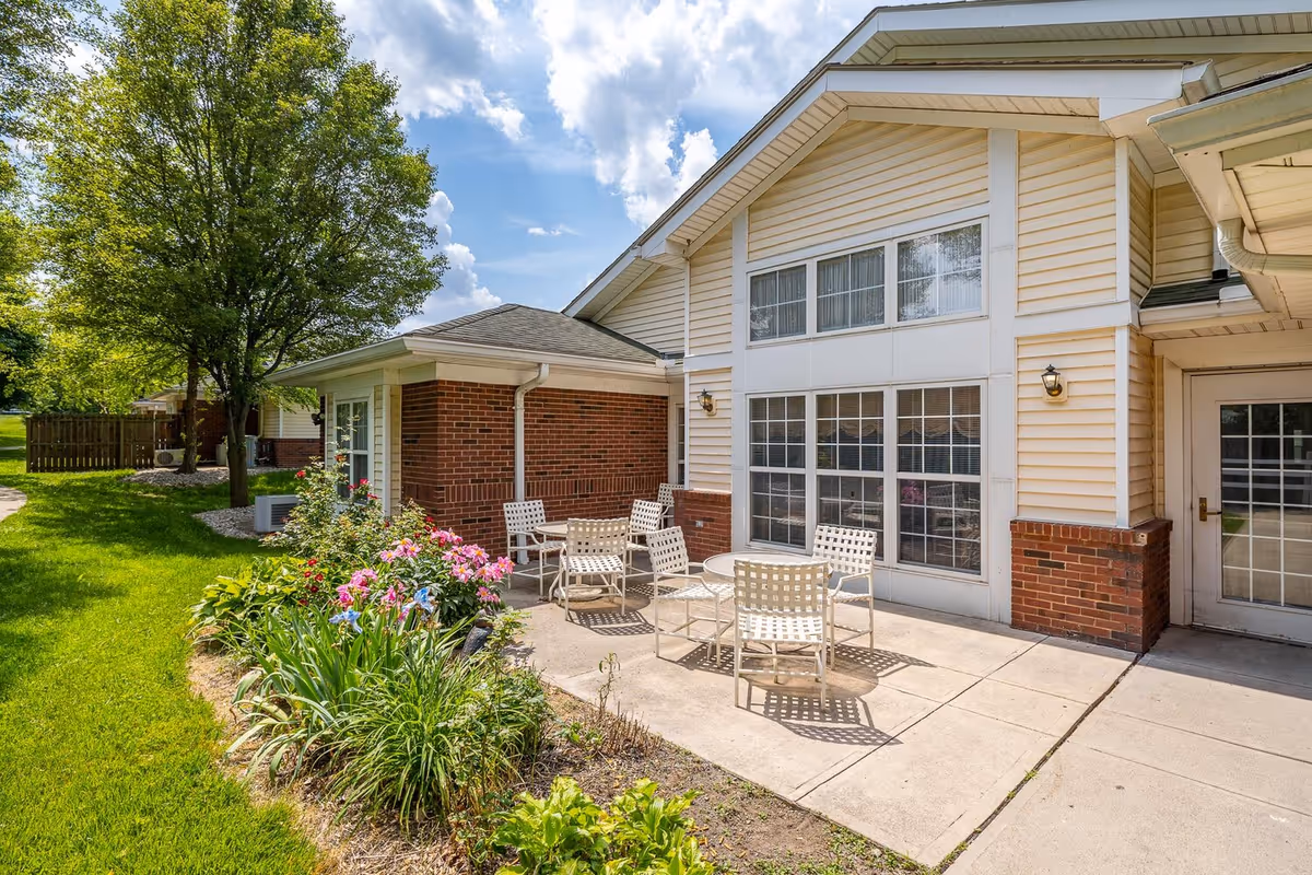 Outdoor patio area at Cottages of Clayton Inc with white metal chairs and tables on a concrete surface, surrounded by green grass, flowering plants, and trees under a partly cloudy sky.