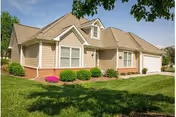 Single-story residential building with beige siding, white trim, and a gabled roof, surrounded by green grass, bushes, and a tree with green leaves in the foreground under a blue sky.