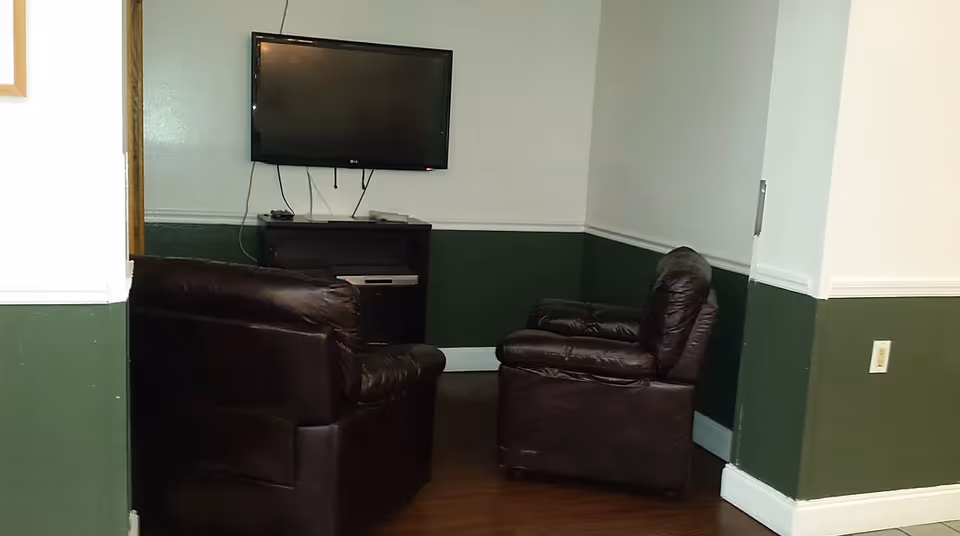 A small lounge with two dark leather armchairs facing a wall-mounted TV above a media stand, with green wainscoting and white upper walls.