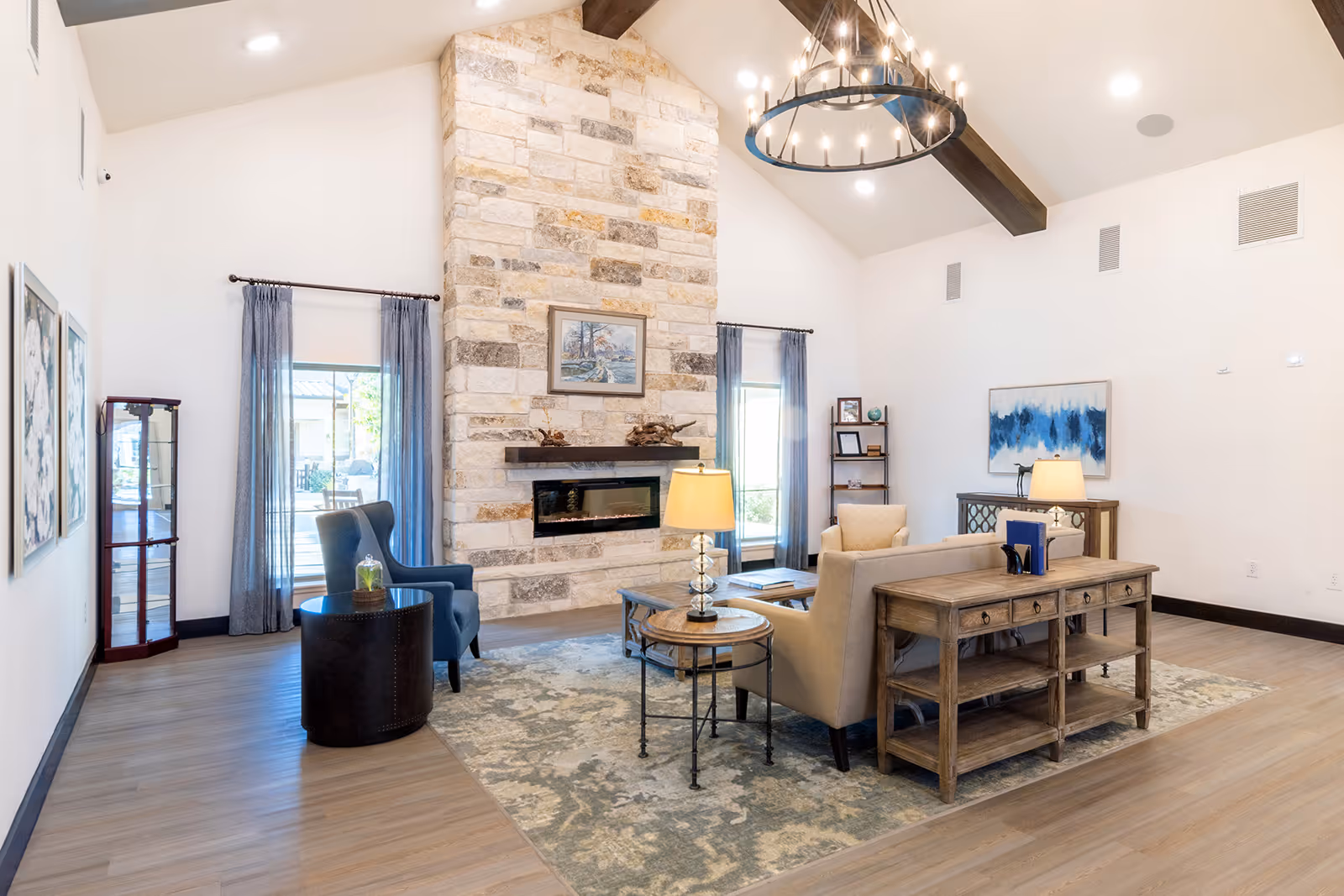 A bright and spacious senior living facility common area with a high vaulted ceiling featuring exposed wooden beams and a large circular chandelier. The room has a stone fireplace centered on one wall with a painting above it. There are two blue armchairs and a beige sofa arranged around a wooden coffee table on a patterned rug. Side tables with lamps flank the sofa. Large windows with sheer curtains allow natural light to fill the room. The flooring is light wood, and there are decorative shelves and artwork on the walls.