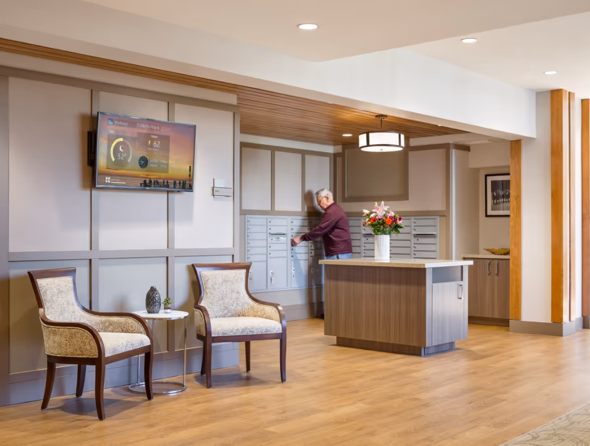 A senior man is retrieving mail from a set of mailboxes in a well-lit, modern lobby area with wooden flooring and neutral-colored walls. Two upholstered chairs with a small round table between them are positioned on the left side, and a flat-screen TV is mounted on the wall above. A wooden island with a vase of flowers is in the center of the room.