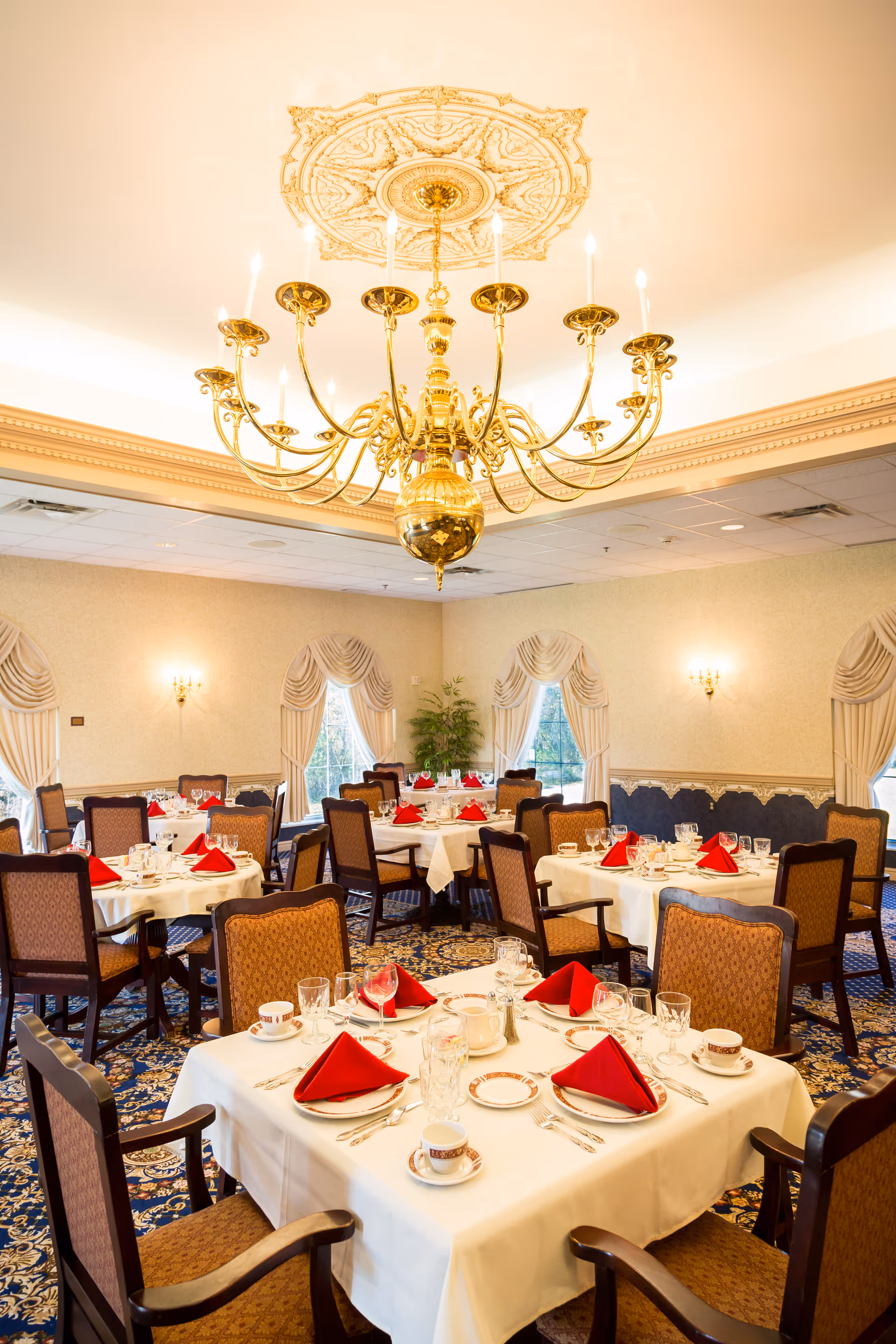 Elegant dining room with round tables covered in white tablecloths, each set with red folded napkins, fine china, glassware, and silverware. The room features ornate curtains on the windows, a large decorative chandelier hanging from the ceiling, and patterned carpet flooring.