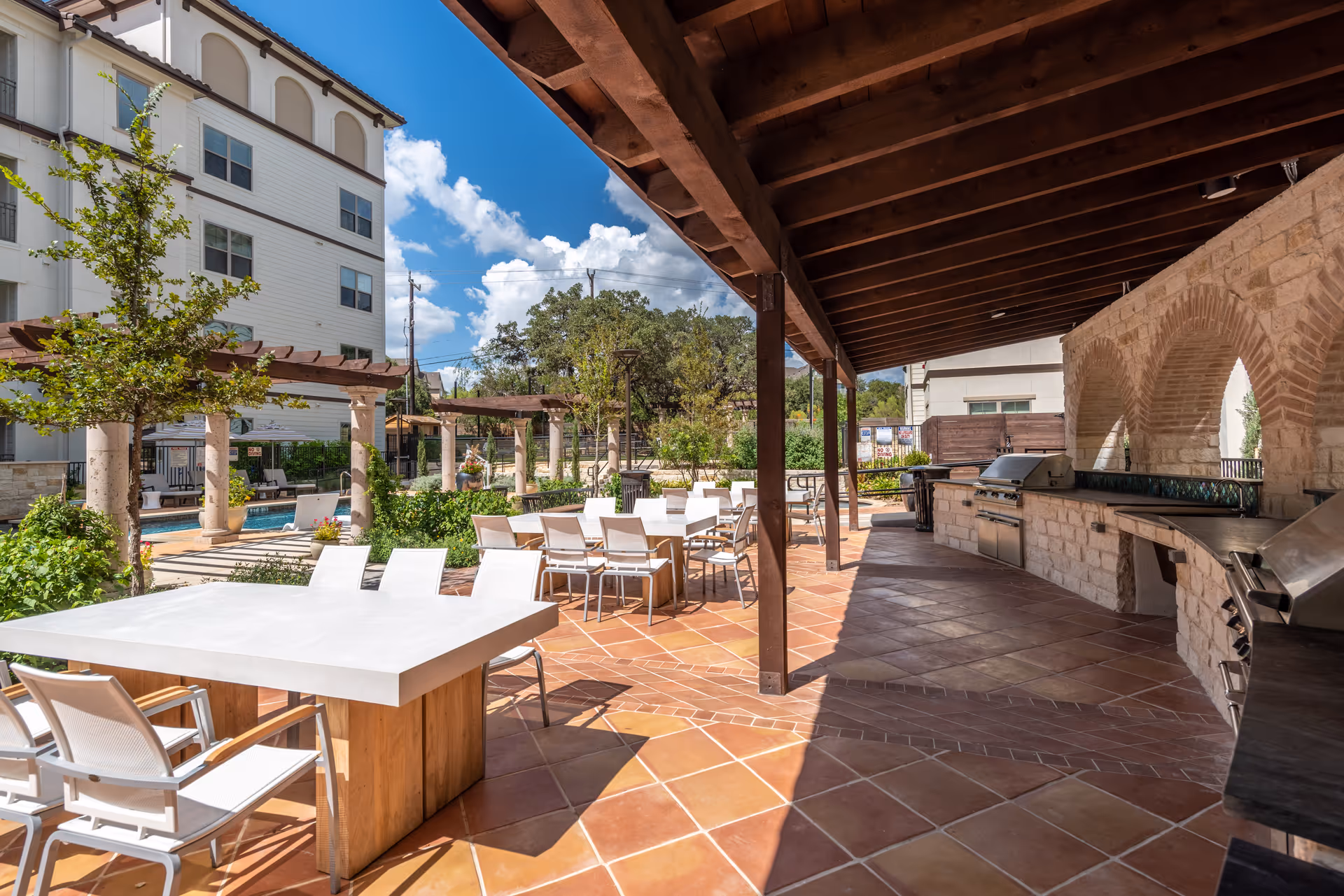 Outdoor patio area at Overture Stone Oak featuring a covered grilling station with multiple grills and countertops, several white tables with chairs, terracotta tiled flooring, pergolas, greenery, and a swimming pool in the background under a blue sky with scattered clouds.
