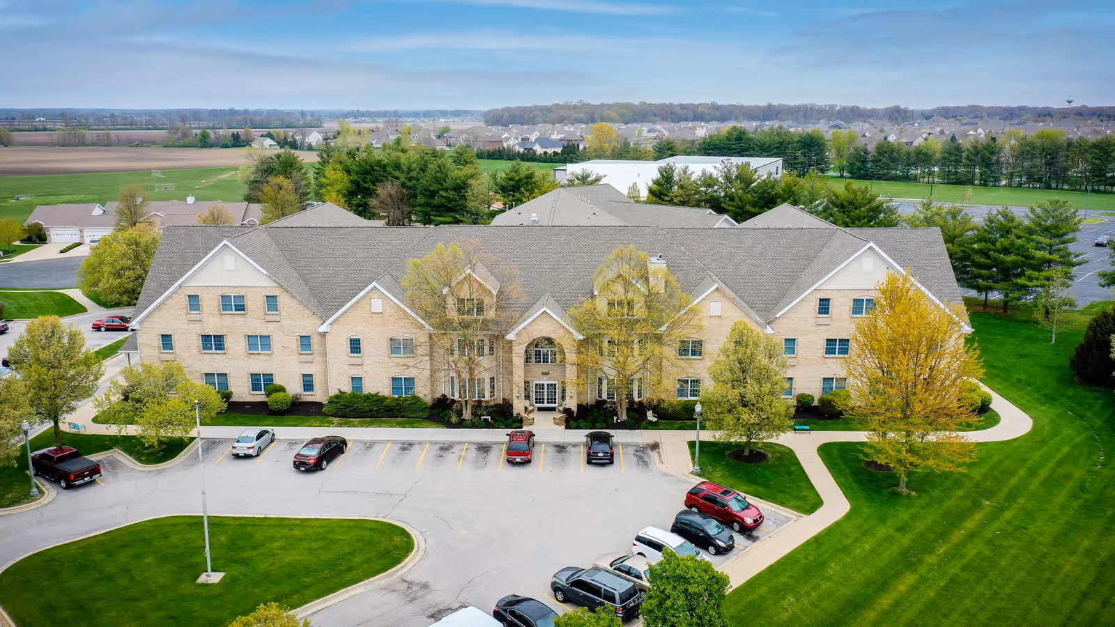 Aerial view of a large, three-story brick building with a gray roof surrounded by green lawns, trees, and a parking lot with several cars parked. The building appears to be a senior living facility with a main entrance featuring large windows and a pathway leading to it. Residential houses and open fields are visible in the background under a partly cloudy sky.