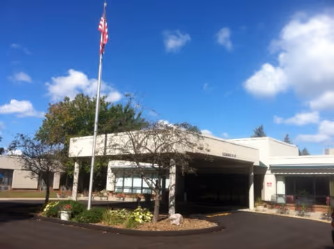 Exterior view of Medilodge of Sterling Heights building under a blue sky with scattered clouds. There is an American flag on a flagpole in front of the building, surrounded by small trees and landscaping. The building has a covered entrance and large windows.
