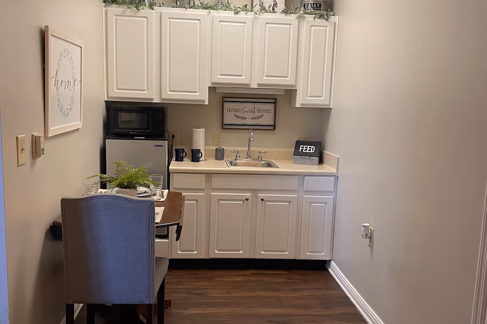 Small kitchenette area with white cabinets, a sink, a microwave on top of a mini refrigerator, and a small wooden table with a chair. The countertop has a paper towel holder, two blue mugs, and a small sign that says 'FEED'. There is a framed 'Home Sweet Home' sign on the wall above the sink and a framed 'home' artwork on the left wall. The floor is dark wood, and there are green plants on top of the cabinets.