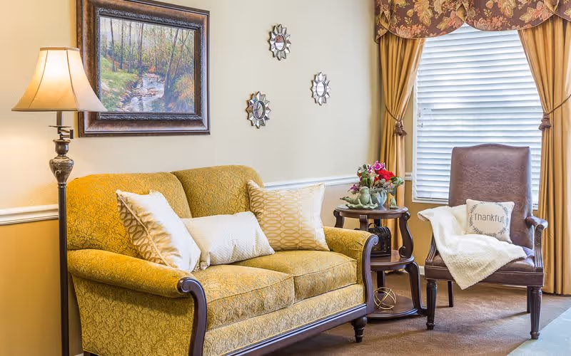 A cozy living room area with a yellow patterned sofa adorned with three decorative pillows, a brown leather armchair with a white throw blanket and a pillow that says 'Thankful', a wooden side table with a flower arrangement, a floor lamp, and a window with closed blinds and floral valance curtains.