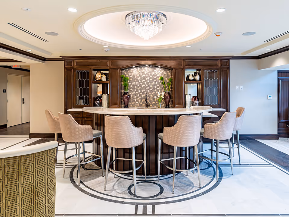 Marble-topped circular bar with six upholstered stools in a well-lit lounge area featuring wood cabinetry and a chandelier.