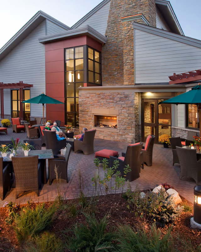 Outdoor patio area at Friendship Village of Bloomington featuring several wicker chairs and tables with green umbrellas. A stone fireplace is built into the building's exterior wall. A few elderly people are seated and conversing near the fireplace. The building has large windows and a combination of stone and siding on the exterior walls. There are plants and landscaping in the foreground.
